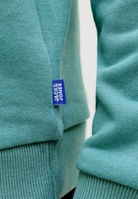 Nærbillede af en sweatshirt i blågrønt strikket stof med ribstrikkede manchetter og et lille blåt mærke med teksten "Jack & Jones" på sidesømmen.