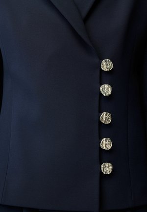 Donkerblauwe blazer met vijf textuurrijke metalen gouden knopen verticaal gerangschikt langs de voorzijde.