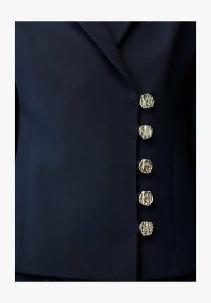 Blazer blu navy scuro con cinque bottoni metallici dorati e lavorati, disposti verticalmente lungo la chiusura frontale.