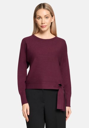 Bordowy sweter z dzianiny z długimi rękawami i wiązaniem w talii. Okrągły dekolt i miękka tekstura. W zestawie z czarnymi spodniami.