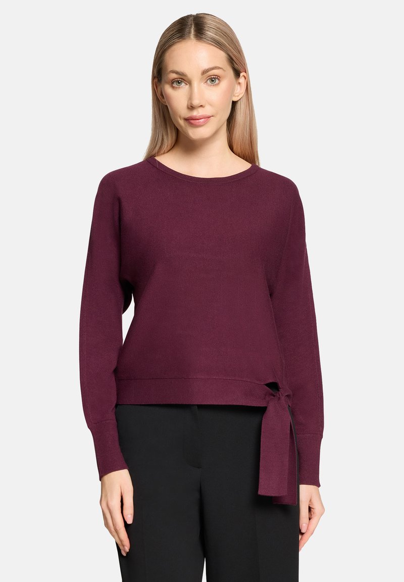 Bordeauxrode gebreide trui met lange mouwen en een strikdetail in de taille. Ronde halslijn en zachte textuur. Gecombineerd met zwarte broek.