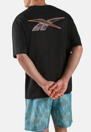 Person mit hinter dem Rücken verschränkten Händen, trägt ein schwarzes T-Shirt mit orangefarbenem Reebok-Logo auf dem Rücken und blaue Shorts mit Camouflage-Muster.