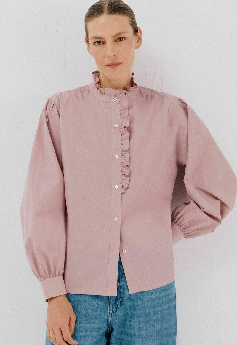 Donna che indossa una blusa rosa chiaro con colletto e parte anteriore arricciati, abbinata a jeans blu, in piedi contro un muro bianco semplice.