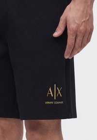 Main droite reposant à côté d'un short noir avec le logo doré "A|X Armani Exchange" près de l'ourlet.