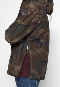 Person iført en grøn og brun camouflagejakke med hætte og synlig sidelukning over en bordeaux skjorte og blå jeans.