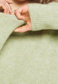 Ljusgrön, mjuk stickad tröja med en texturerad, något ludden finish. Ärmarna är uppviktade, vilket visar händerna mot en matchande bakgrund.