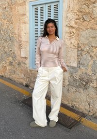 Gestreiftes Langarm-Polo in sanftem Beige und Weiß, kombiniert mit locker sitzenden weißen Hosen. Graue Slipper. Der Hintergrund zeigt eine strukturierte Steinmauer.