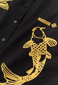Tissu noir avec des poissons koi brodés en jaune et le mot « WOODBIRD » près de boutons noirs.