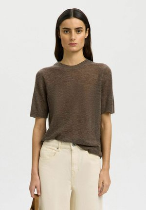 Žena s dlhými tmavými vlasmi má na sebe hnedý krátky sveter a krémové nohavice s vysokým pásom, stojaca pred svetlým jednofarebným pozadím.