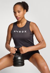 Schwarzes Sporttanktop mit "HYROX"-Logo, kombiniert mit einer schwarzen Kettlebell in den Händen. Hautton ist medium; auffällige Tattoos an den Armen.