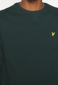Sudadera de color verde oscuro con cuello redondo, ribete en el cuello y un pequeño logotipo de un pájaro amarillo en el lado izquierdo del pecho. Textura suave, diseño casual.