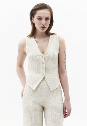 Femme aux cheveux châtain clair portant un gilet sans manches crème texturé et un pantalon taille haute assorti, debout devant un fond blanc.