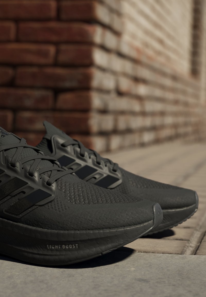 Pár černých běžeckých bot Adidas s podrážkami Light Boost umístěný na dlažebním povrchu venku poblíž cihlové zdi.
