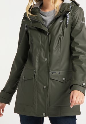 Femme portant une veste imperméable vert foncé avec capuche, boutons-pression, poches avant et cordons à motifs sur une chemise grise.