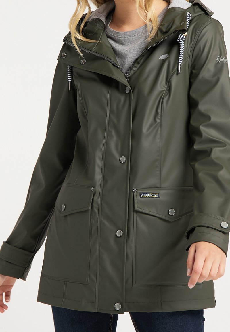 Femme portant une veste imperméable vert foncé avec capuche, boutons-pression, poches avant et cordons à motifs sur une chemise grise.