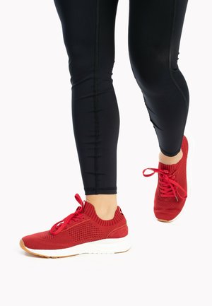 Rote gestrickte Sneakers mit atmungsaktivem Design, ausgestattet mit einem gerippten Knöchelbund und weißer Sohle, kombiniert mit schwarzen Leggings.
