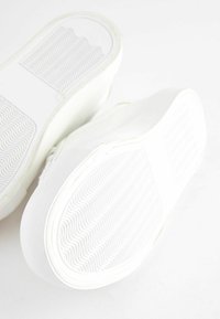 Baskets blanches avec semelles en caoutchouc, présentant un motif de grip texturé et des bords lisses et arrondis. La tige est également blanche et minimaliste.