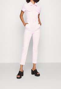 Rosa denimoverall med korta ärmar, två bröstfickor och midja med bälte. Matchat med svarta platåsandaler.