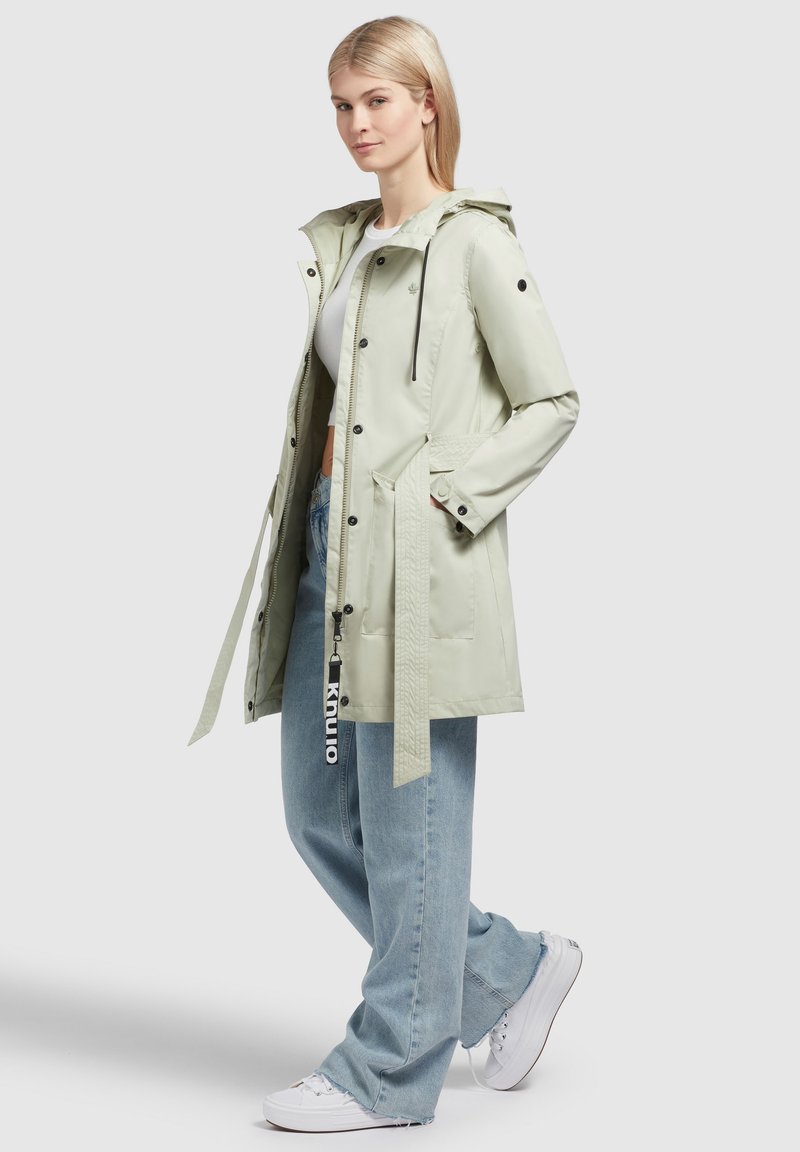 Giacca impermeabile verde chiaro con cappuccio, bottoni a pressione, tasche laterali, cintura regolabile in vita e dettaglio intrecciato sulla parte anteriore.
