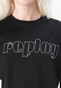 Juodas medvilninis marškinėliai su baltais siuvinėjimais tekstu "replay" stilizuotu šriftu, apvaliu iškirpimu ir trumpomis rankovėmis.