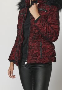 Kobieta nosząca czerwoną i czarną kurtkę puchową w wzory z futrzanym kapturem, w czarnym topie i czarnych skórzanych spodniach, poprawiająca kołnierz kurtki.