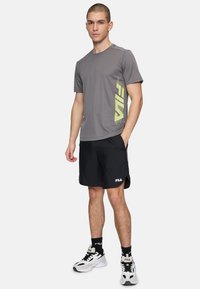 Camiseta atlética gris con logo amarillo, combinada con pantalones cortos negros y zapatillas blancas. El conjunto presenta un ajuste moderno y tejido transpirable.