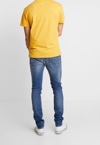 Žluté bavlněné tričko s krátkým rukávem v kombinaci s modrými úzkými džíny a bílými teniskami, pohled zezadu.