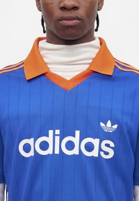 Νέος με πλεξούδες που φορά μπλε μπλούζα Adidas με πορτοκαλί γιακά και λευκή ζιβάγκο από μέσα, χωρίς εμφανές φόντο.