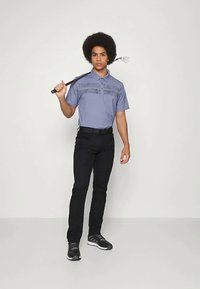 Polo majica s kratkimi rokavi v vijolični barvi s horizontalnimi črtami na prsih, v kombinaciji s črnimi hlačami in črnimi golf čevlji, drži golf palico.