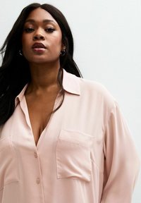 Blusa rosa chiaro realizzata in tessuto morbido, con chiusura a bottoni, collare appuntito e due tasche sul petto. Design morbido e drappeggiato con maniche lunghe.
