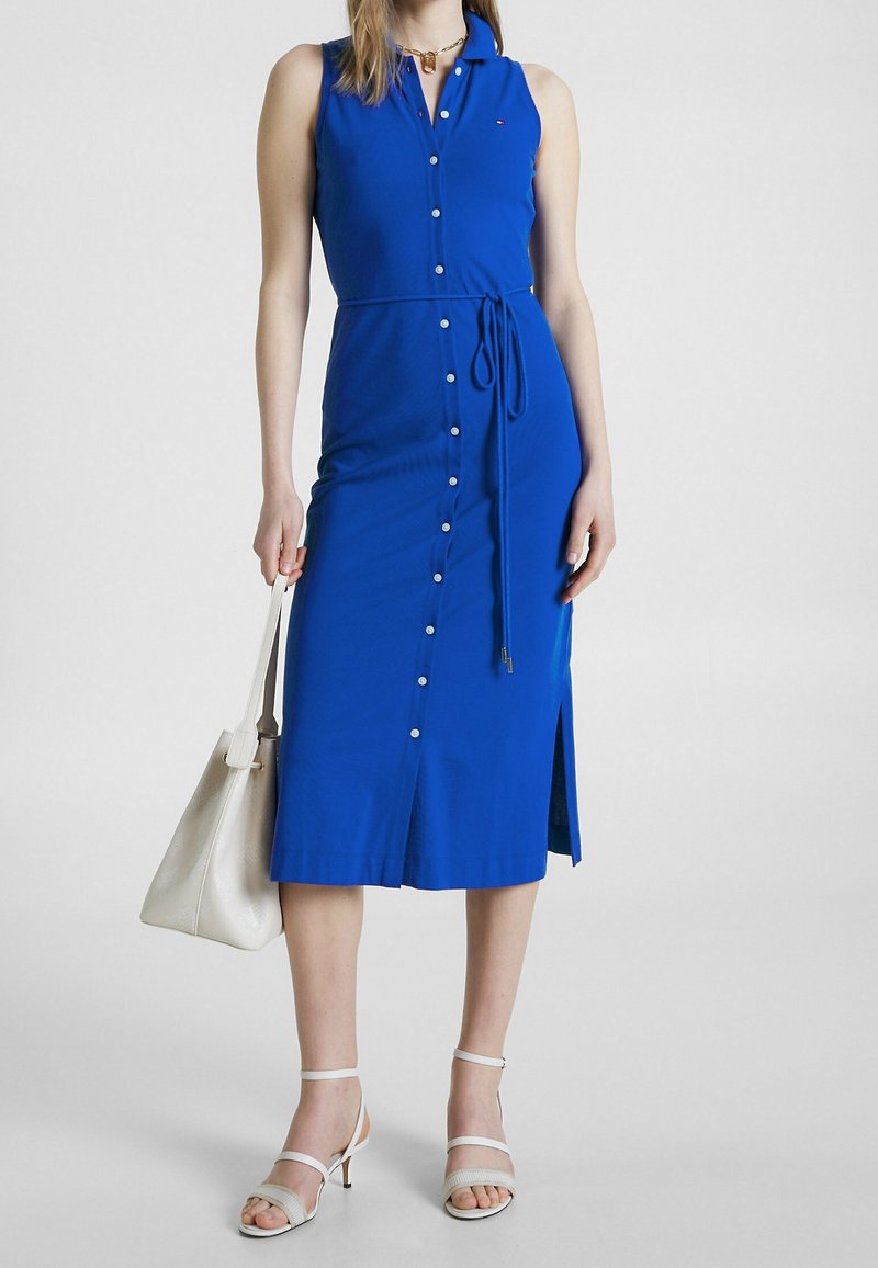 Blå, ärmlös klänning med knappstängning fram, midjeband, slits på sidan och mjukt tyg. Bärs med vita sandaler med remmar och en lätt handväska.