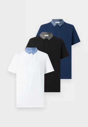 Homme portant un polo blanc avec un col bleu, un pantalon bleu foncé, des chaussures noires et une montre-bracelet, debout devant un fond blanc.
