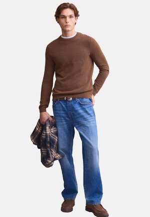 Giovane uomo con maglione marrone, jeans blu e scarpe marroni, che tiene una giacca fantasia nella mano sinistra.