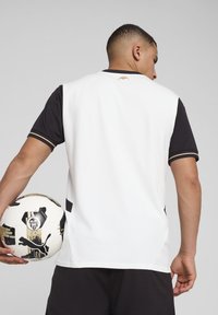 Valge ja must spordijersey, lühikeste varrukatega, millel on kuldsed aktsendid ja logo, mille hoiab käes inimene koos mustrilise jalgpalliga.