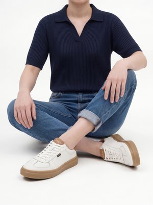 Personne assise en tailleur portant un haut côtelé bleu marine à manches courtes, un jean bleu retroussé et des baskets blanches avec des semelles marron et la marque "Replay".