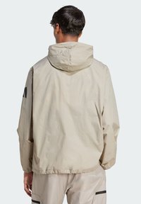 Light beige hooded jacket with a loose fit, elastic hem, and contrasting black brand logo on the left sleeve. Smooth, slightly crinkled texture.