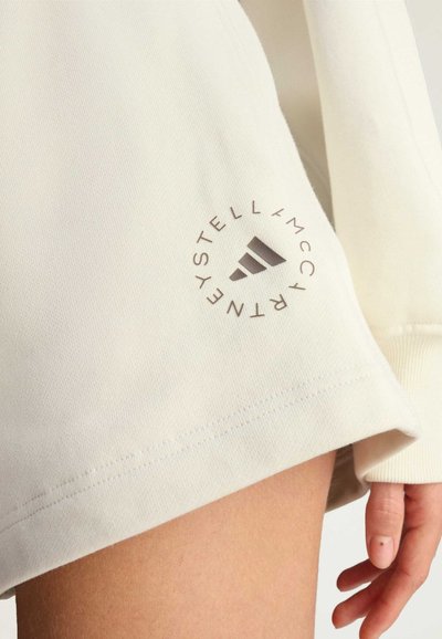 Cremefarbenes, locker sitzendes Sweatshirt mit einem runden Logo, das Streifen und Text in der Nähe des Saums zeigt und aus weichem, strukturiertem Stoff besteht.
