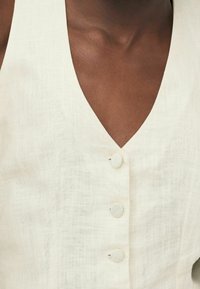 Witte linnen top met een diepe V-hals, voorzien van drie met stof bedekte knopen aan de voorkant. De textuur lijkt licht en enigszins gestructureerd.