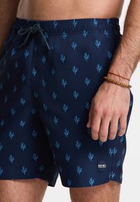 Tengerész kék fürdőshorts kék hímzett korall mintázattal. Rugalmas derékpánt zsinórral, oldalsó zseb és SHIWI logós badge a szegélyen.
