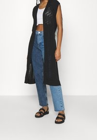 Chaleco largo negro con frente abierto, con patrones texturizados y diseño sin mangas, combinado con una blusa blanca corta, jeans azules y sandalias negras.
