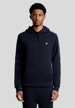 Jeune homme portant un sweat à capuche noir avec un logo d'oiseau jaune sur la poitrine et des bandes de marque le long des manches, debout devant un fond gris.