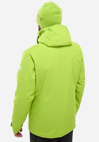 Veste imperméable vert vif avec capuche, présentant une texture lisse et un logo sur la manche gauche. Pantalons noirs visibles en dessous.