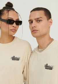 Two models wearing beige "Rome" logo t-shirts; one with black sunglasses and hair in buns, the other with short hair and a silver chain necklace.