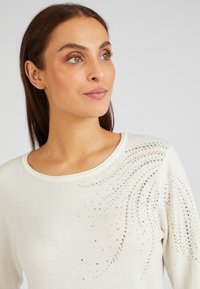 Witte gebreide trui met een ronde halslijn en versieringen van kleine gouden, zilveren en heldere rhinestones in een spiraalpatroon.