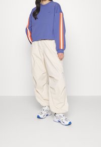 Persona che indossa un top a maniche lunghe viola con strisce arancioni e bianche sulle maniche, pantaloni larghi color crema e sneakers bianche New Balance con suole blu.