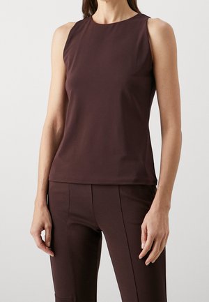 Femme portant un top marron foncé ajusté sans manches et un pantalon assorti, debout devant un fond clair uni.
