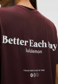 Bordó póló fehér felirattal: "Jobb minden egyes nap", "lululemon" és "Train Crew Class of 1998", amelyet egy személy visel hátulról látható.
