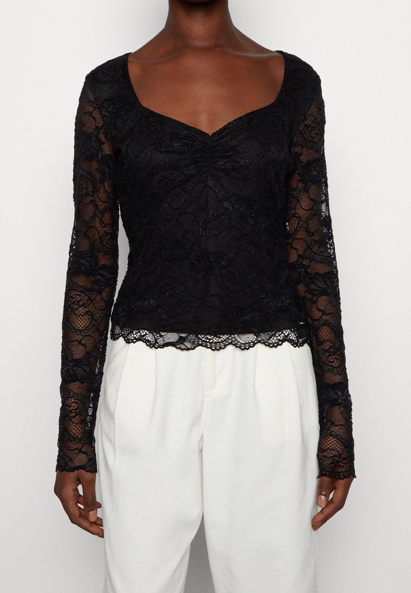 Femme portant un haut en dentelle noire à manches longues avec des bords festonnés, associé à un pantalon blanc taille haute sur un fond uni.
