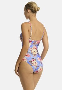 Vrouw in een bloemenbadpak, met haar rug naar de camera gekeerd, tegen een blauwe achtergrond met roze en gele bloemen. Haar haar in een bun gestyled, elegante pose.