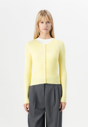 Ribbet, lys gul cardigan med lange ermer og rund hals, med frontknapp-lukking og en kortere kant. Kombinert med grå bukser.